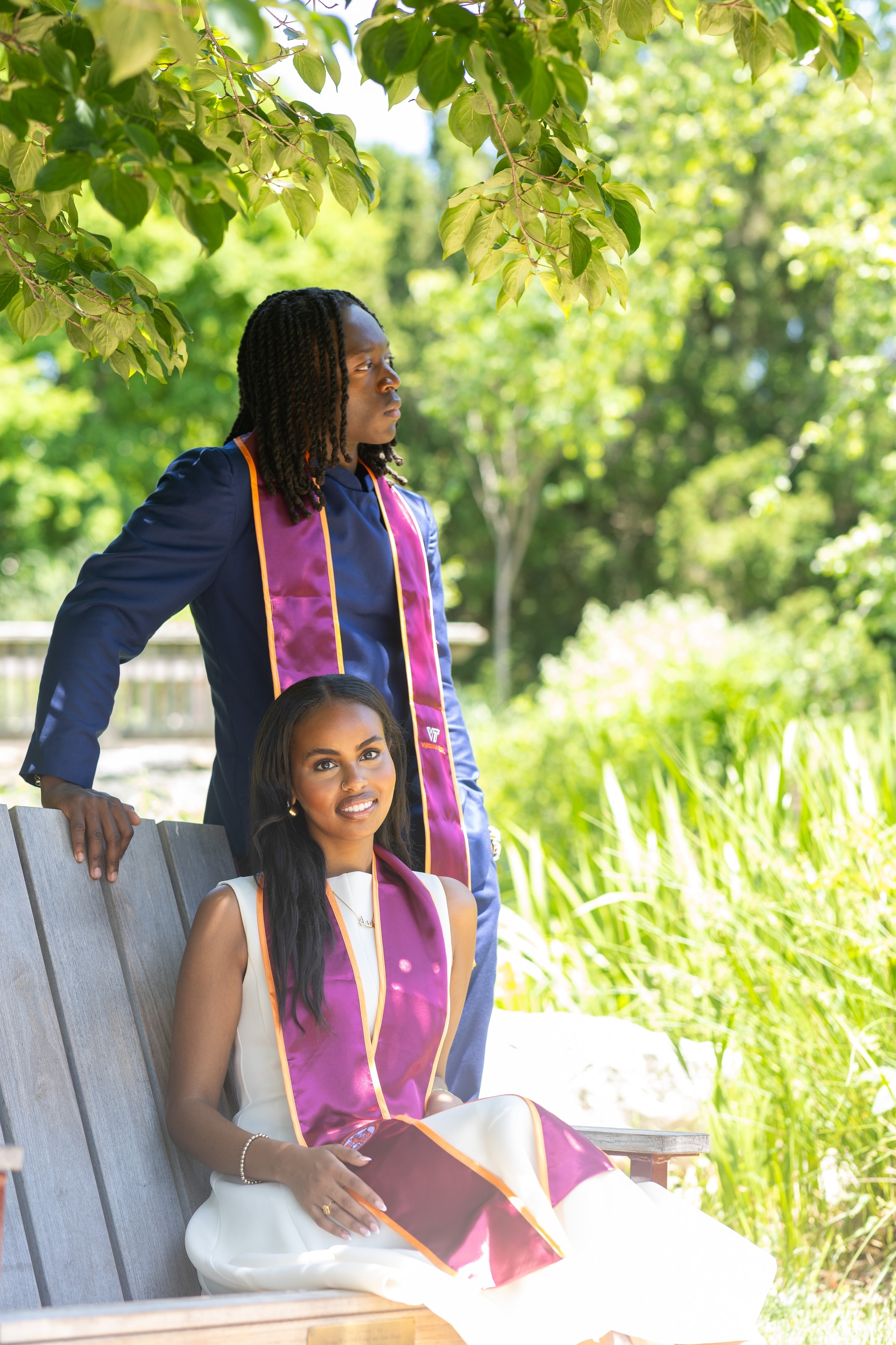 Two graduates wearing purple graduation stoles outdoors under a tree, one standing behind a bench and one seated in front, smiling