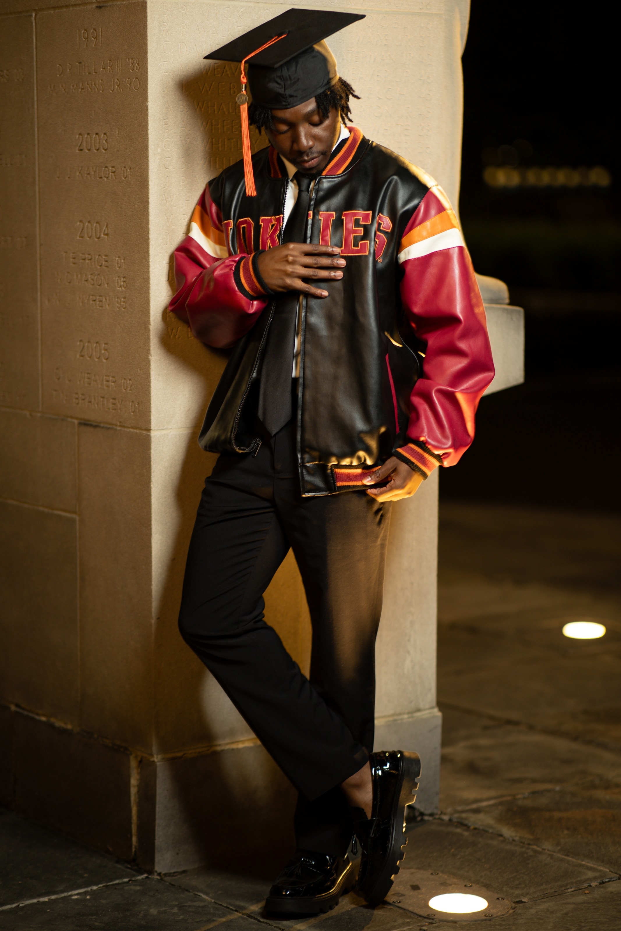 A graduate in a cap with an orange tassel and a colorful varsity jacket, leaning against a stone pillar at night, eyes closed