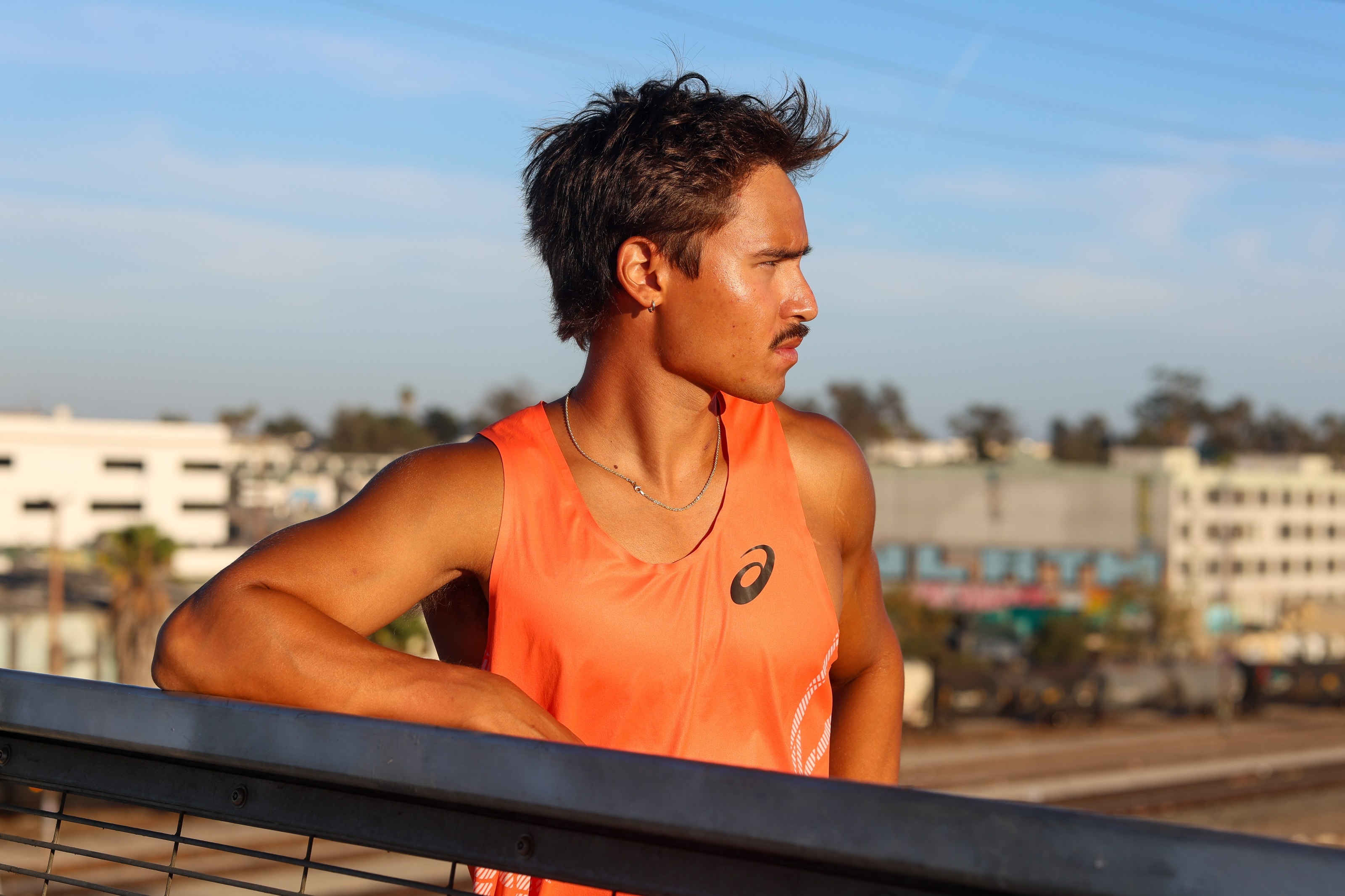 Christian leaning against a bridge railing at golden hour, looking off to the side in profile, city buildings blurred behind him