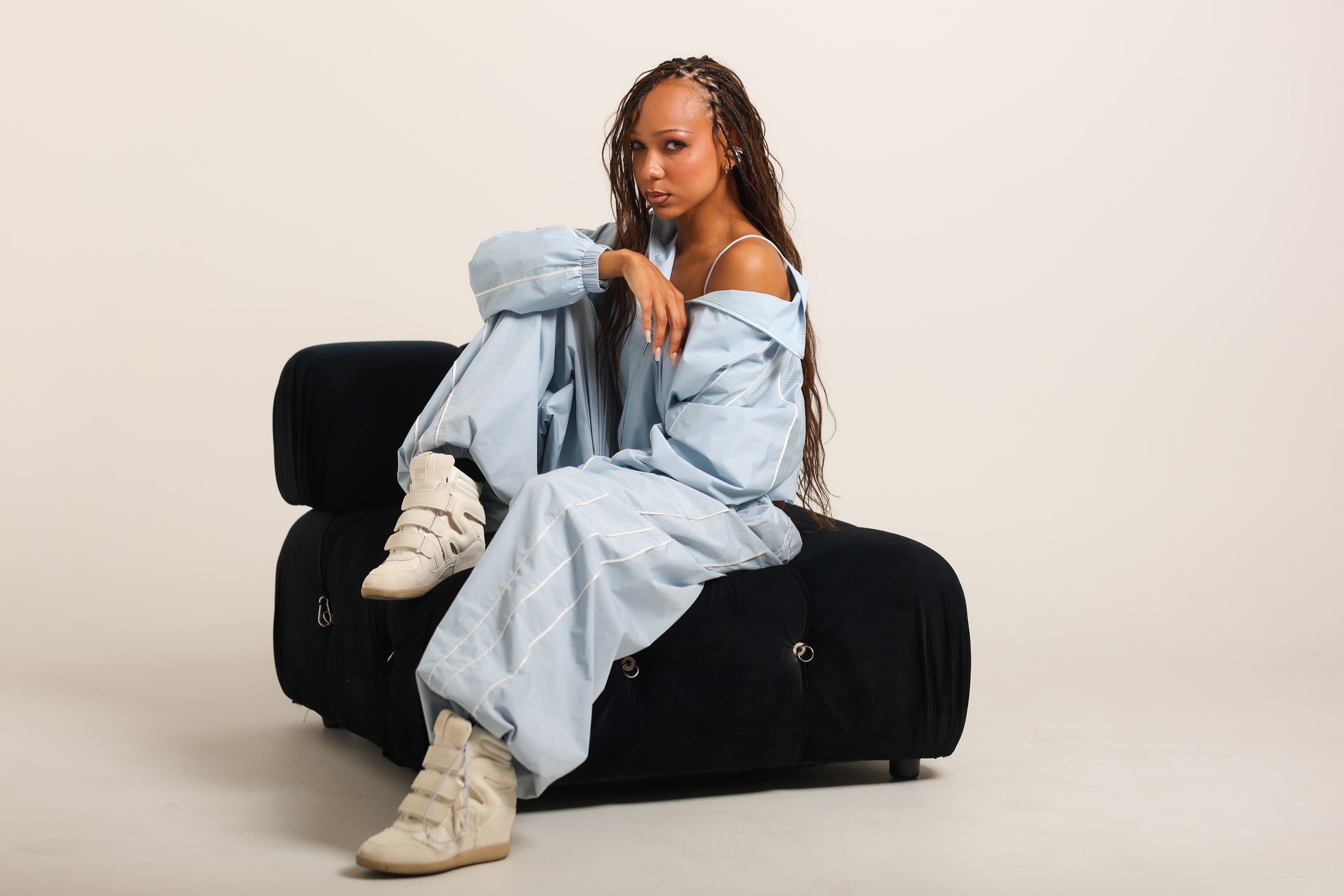 Ciara sitting on a black tufted bench in an oversized sky-blue tracksuit, one arm resting on her knee, looking directly into the lens