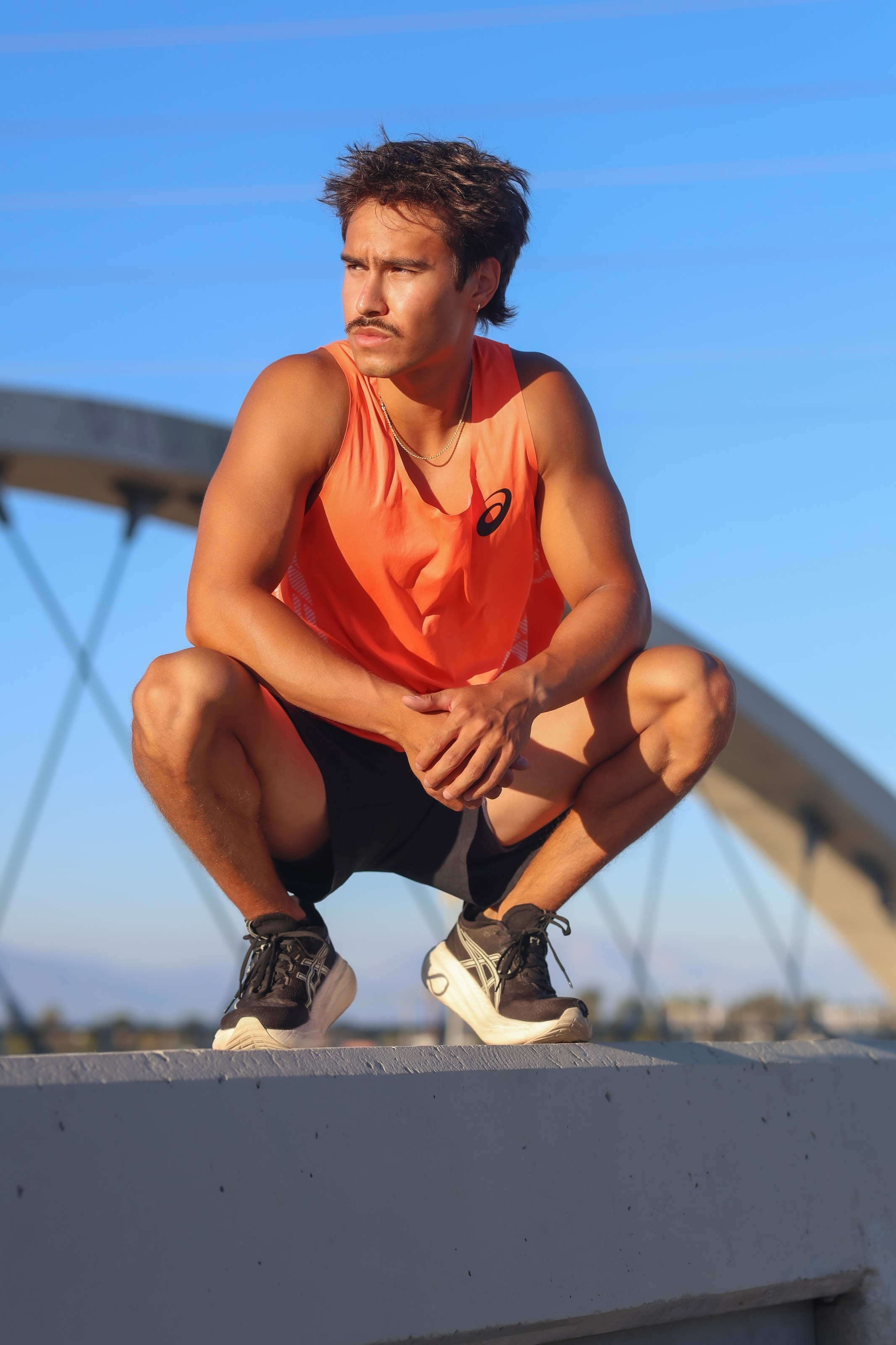 Christian crouched on a concrete ledge in an orange tank top, a steel bridge arch behind him