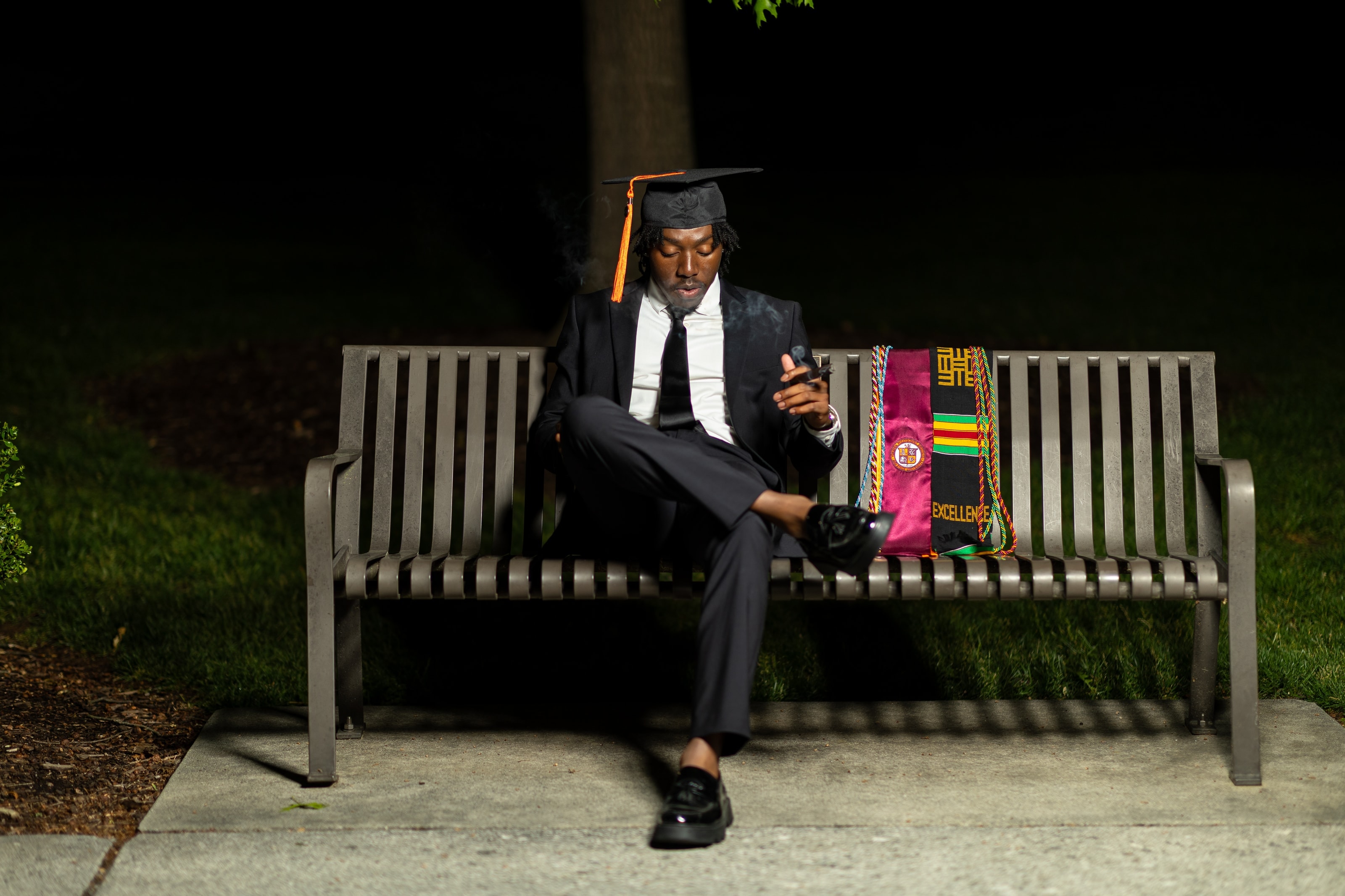 Ethan sitting on a park bench at night in a cap and gown stole, smoking a cigar with his graduation regalia beside him