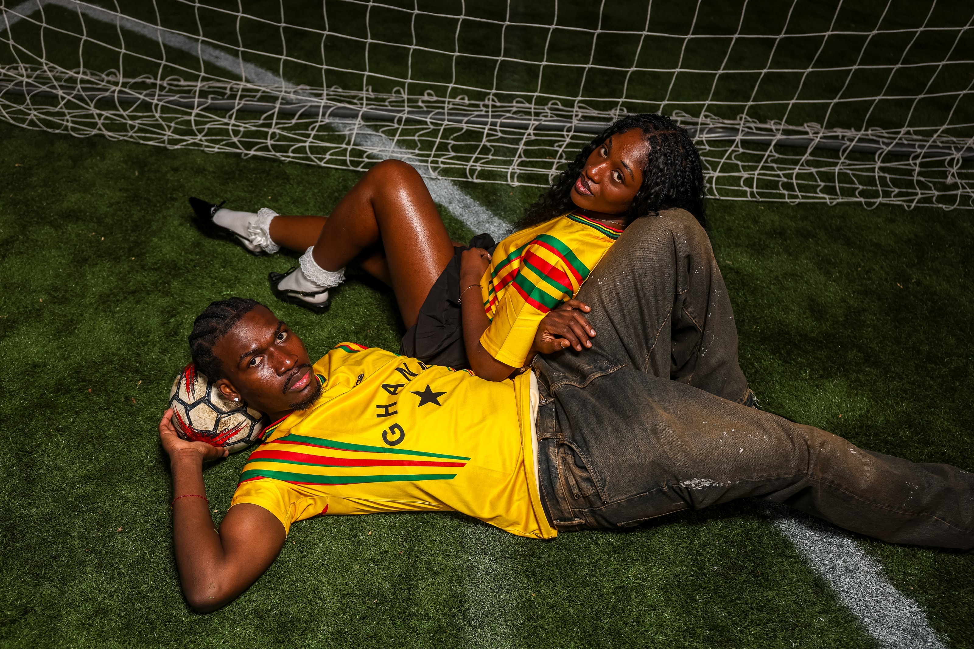 A couple lying together on the turf inside a soccer goal at night, both in yellow Ghana jerseys, looking directly into the lens