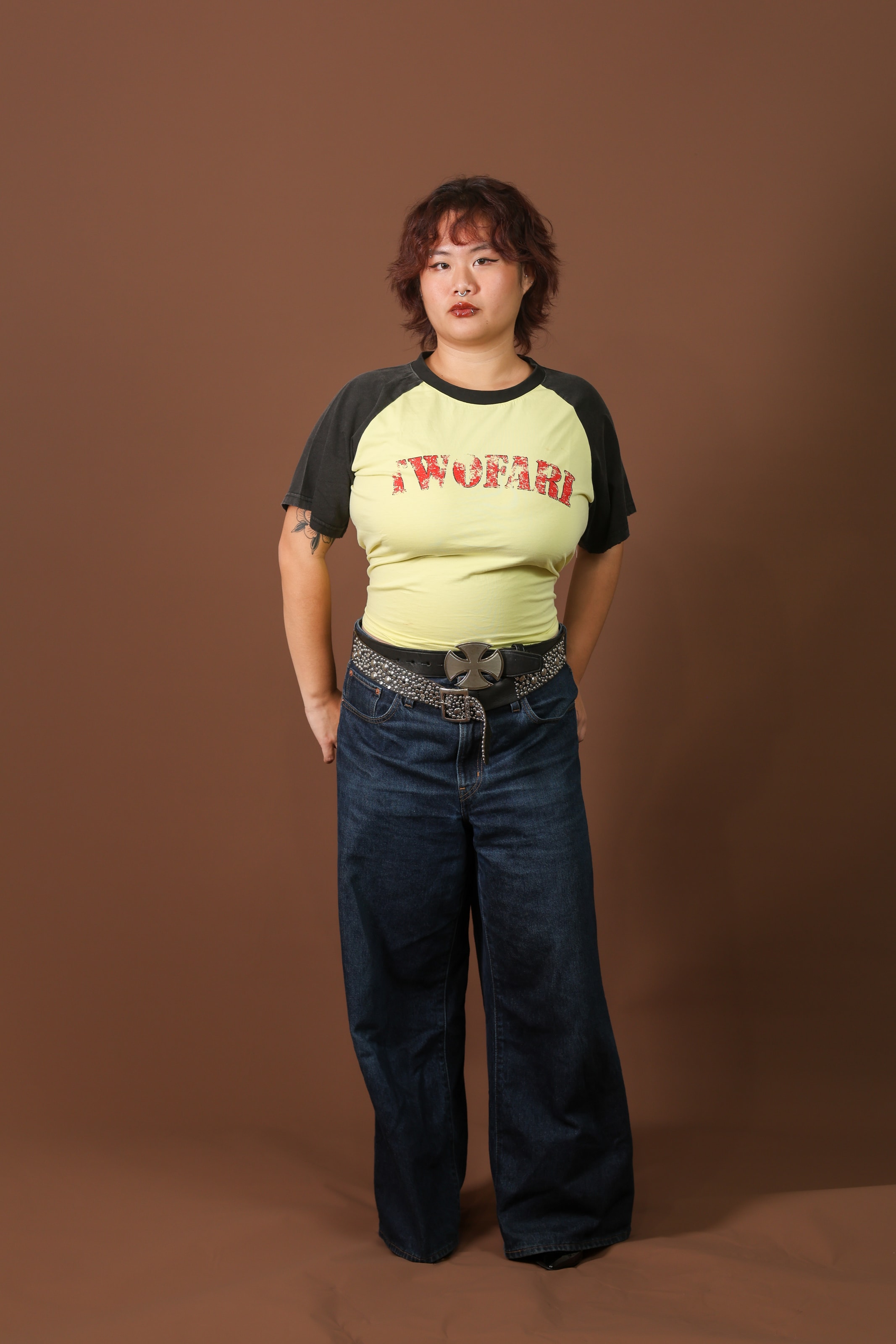 A woman in a TWOFARI tee and studded belt, hands clasped behind her back, looking off to the side
